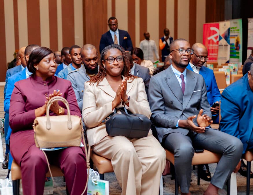 Une photo capturant un moment de la conférence de Bamboo Microfinance à Brazzaville le 15 septembre 2026. L'assistance est composée d'hommes et de femmes en tenue professionnelle (costumes, vestes), assis sur des chaises de conférence. Au premier plan, une femme en veste beige et un homme en veste grise sont assis à une table, écoutant attentivement l'intervenant. Plusieurs autres personnes sont visibles derrière eux, certaines en uniformes bleus. L'arrière-plan montre des rideaux à rayures et une bannière de l'événement.