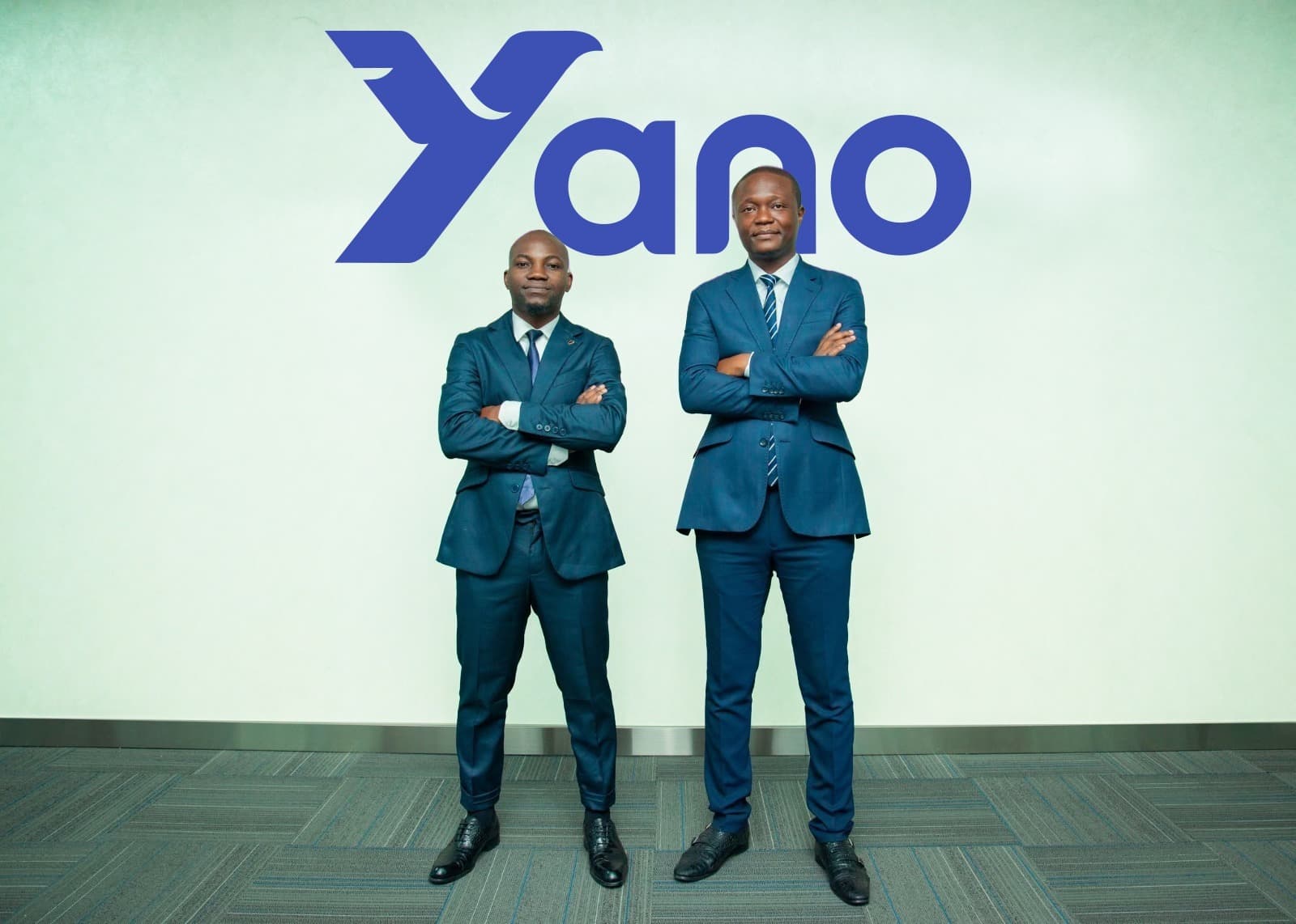 Deux hommes d'affaires d'origine africaine sont debout côte à côte, bras croisés et souriant légèrement devant un mur blanc orné du logo bleu vif de "Yano". Ils portent tous deux des costumes bleu marine bien ajustés, des chemises blanches et des cravates assorties. L'homme de gauche (Régis Dilou) est un peu plus petit, avec une coupe rase et une barbe légère, tandis que l'homme de droite (Elwin Gomo) est plus grand et porte une coiffure afro courte. Le sol est un tapis à motifs géométriques gris. L'éclairage est clair et uniforme, provenant probablement du plafond d'un bureau moderne.
