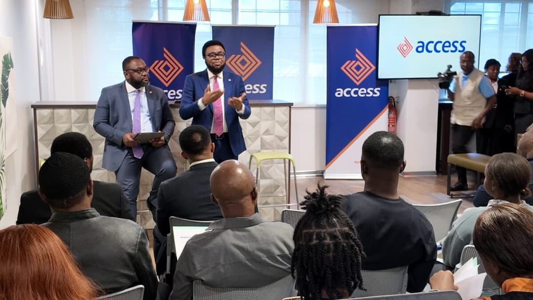 Cette image capture un moment clé du lancement d'Access Invest. Un orateur d'Access Bank, portant un costume bleu et une cravate rose, parle de manière dynamique, les mains gesticulant pour appuyer ses propos. Il est flanqué d'un autre cadre de la banque assis sur un tabouret, et derrière eux se trouvent des bannières Access Bank et un écran de télévision affichant le logo de la banque. Un caméraman est visible en arrière-plan, capturant l'événement. L'image est prise depuis le public, montrant l'arrière des têtes des participants au premier plan.