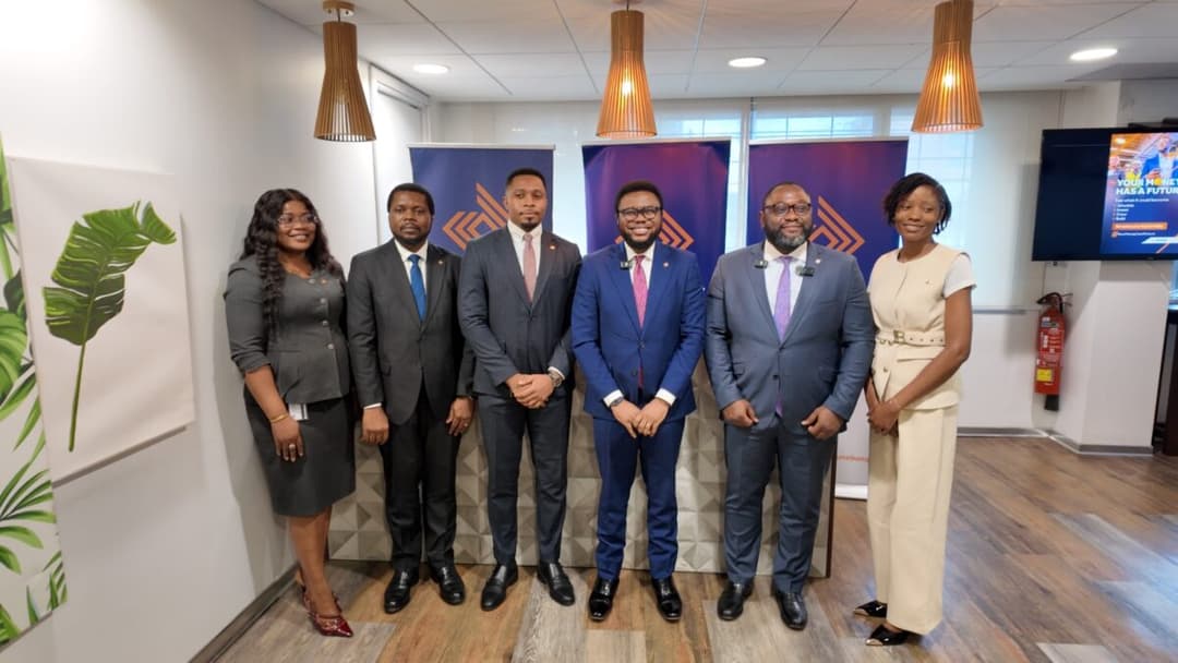 Une photo de groupe réunissant six cadres d'Access Bank Cameroon, dont quatre hommes et deux femmes, tous vêtus de tenues professionnelles élégantes. Ils se tiennent debout et sourient devant des bannières violettes affichant le logo d'Access Bank, dans une salle moderne bien éclairée par des suspensions en bois et des lumières encastrées.