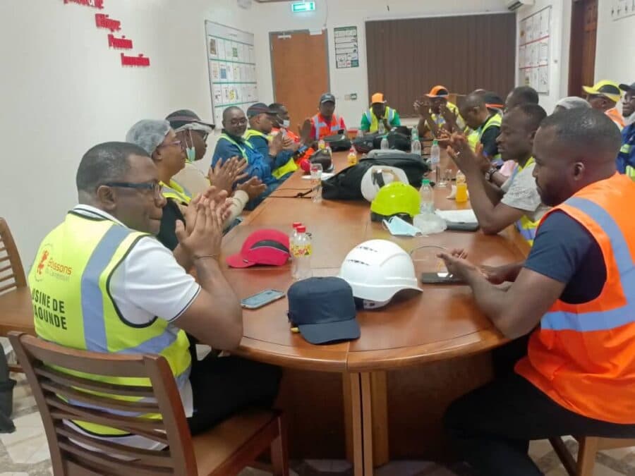 Une photo de groupe prise lors d'une réunion technique dans une salle de conférence à la Castel Academy, où des professionnels de Boissons du Cameroun, dont un homme au premier plan avec un gilet haute visibilité portant le logo de l'entreprise et la mention "USINE DE DOUALA", discutent et applaudissent. L'image capture l'atmosphère de collaboration et de planification du programme « Zéro Défaut Conformité Food Safety » pour standardiser les pratiques de sécurité sanitaire.