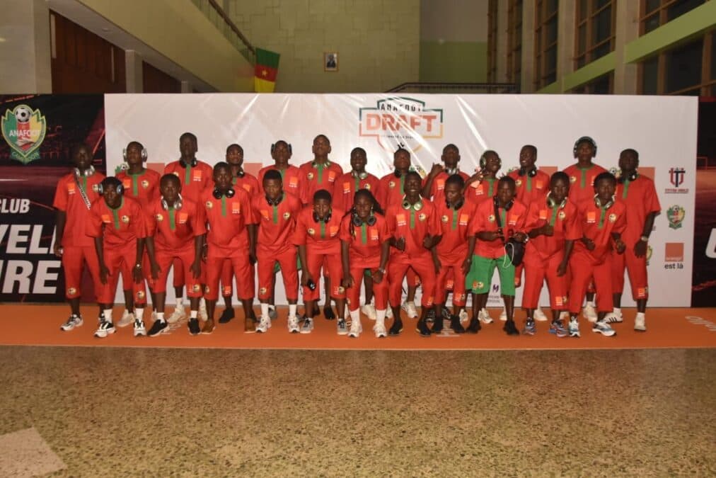 Une photo de groupe alignée de dizaines de jeunes footballeurs, tous portant une tenue rouge unifiée (maillot et short) et, pour la plupart, des écouteurs blancs autour du cou. Ils sont positionnés sur un tapis orange et posent avec assurance devant un grand mur d'arrière-plan (backdrop) rempli de logos répétitifs d'Orange, d'ANAFOOT, et de la bannière "ANAFOOT DRAFT". Le cadre suggère un couloir spacieux ou une zone de réception, capturant l'enthousiasme et l'unité de cette nouvelle cohorte de professionnels.