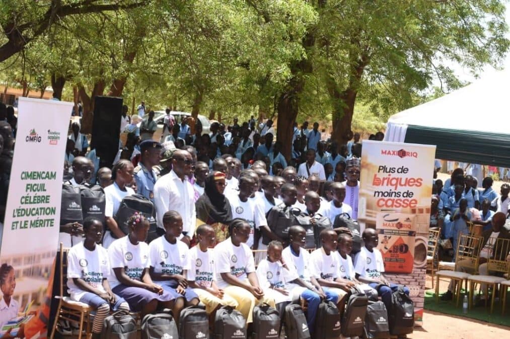 Vue d'ensemble de la cérémonie de remise des kits scolaires à Figuil. Les élèves bénéficiaires sont assis au premier rang avec leurs kits scolaires posés devant eux, entourés par les membres de la communauté, les représentants de CIMENCAM, les forces de l'ordre et les autorités administratives sous des bannières promotionnelles de l'entreprise.