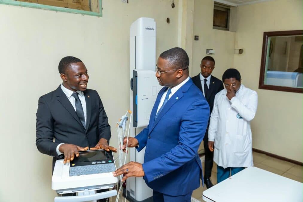 Un spécialiste technique de Gima-Bambot MedTech explique le fonctionnement d'une console médicale à écran tactile à un cadre dirigeant de BGFIBank vêtu d'un costume bleu. À l'arrière-plan, un médecin en blouse blanche et d'autres collaborateurs observent la démonstration devant un système de radiographie numérique vertical Mindray.
