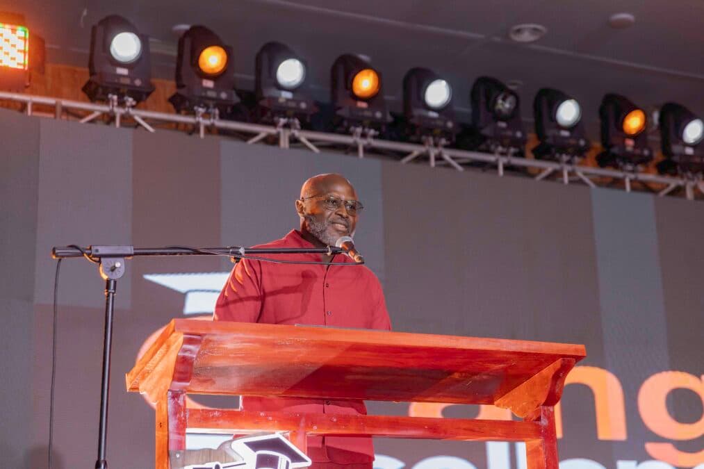 Photo de Patrick Benon, DG d'Orange Cameroun, portant des lunettes et une chemise rouge, debout derrière un pupitre en bois. Il s'exprime dans un microphone lors de la cérémonie Orange Excellence+ 2026. En arrière-plan, on aperçoit des éclairages de scène et une partie du logo "Orange Excellence".