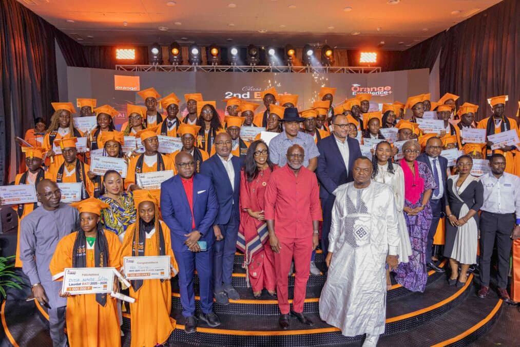 Photo de groupe plus large sur scène, incluant Patrick Benon, DG d'Orange Cameroun (en rouge), des officiels et partenaires, ainsi que les 50 lauréats en toges orange. Certains lauréats tiennent des chèques géants. En arrière-plan, on voit l'inscription "Orange Excellence+ 2026 - Class of 2nd Edition" et une partie du logo Orange.