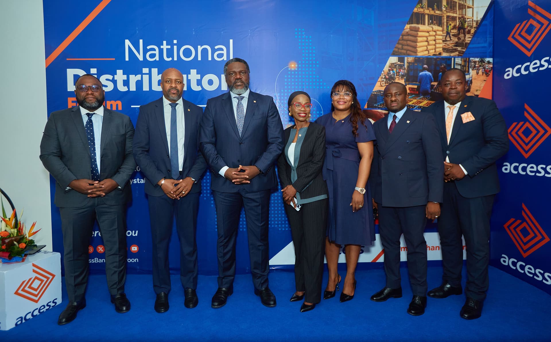 Une photo de groupe posée en intérieur montrant sept cadres de haut niveau d'Access Bank Cameroon, en tenue professionnelle élégante (costumes sombres pour les hommes, robes et tailleur pour les femmes). Ils sont alignés devant une toile de fond d'événement de couleur bleu roi et orange portant l'inscription lisible « National Distributors Forum » et le logo de la banque. M. Irechukwu est au centre, les mains jointes.