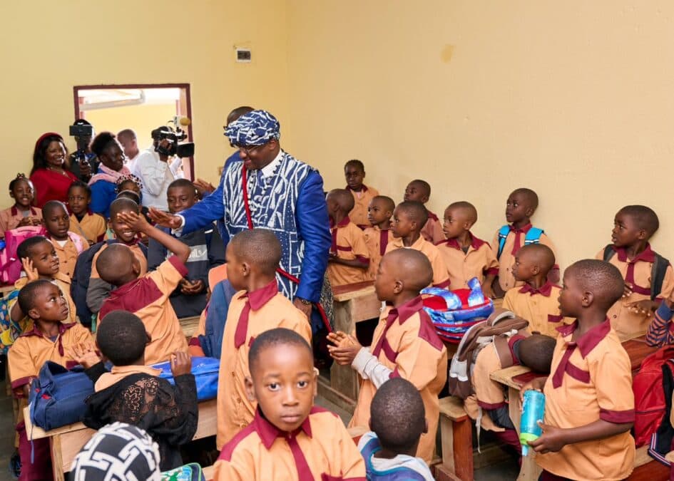 L'image montre le Représentant de la Fondation, Abakal Mahamat, en tenue d'apparat, souriant et échangeant avec de nombreux élèves portant leur uniforme scolaire (beige et marron). Ils sont dans une salle de classe propre, aux murs repeints, où l'on voit les nouveaux bancs et les fournitures.