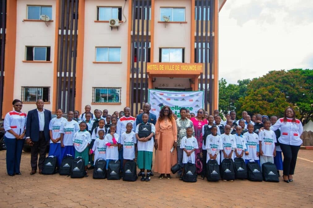 Photo de groupe réunissant les élèves lauréats du programme « Excellence Scolaire » initié par CIMENCAM, accompagnés des autorités locales et des responsables d'entreprise. Les élèves, vêtus de t-shirts blancs promotionnels, posent avec leurs nouveaux sacs à dos scolaires posés au sol devant le bâtiment de l'Hôtel de ville de Yaoundé IIIe.