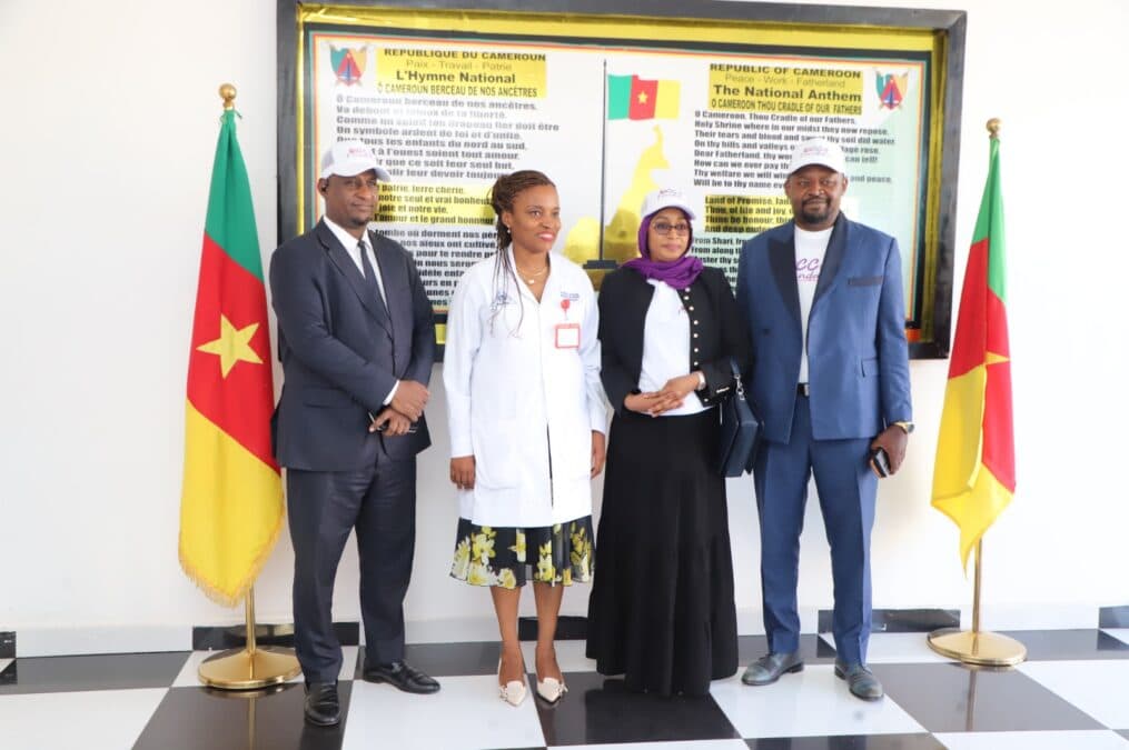 Pose officielle de quatre responsables de CCA-BANK et du corps médical, encadrés par deux drapeaux du Cameroun et un tableau mural de l'hymne national dans le hall de réception de l'hôpital.