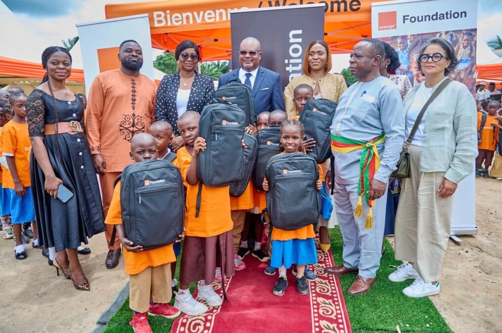 Une photo de groupe posée lors de la cérémonie officielle. Au centre, des élèves en t-shirts orange tiennent fièrement leurs nouveaux cartables noirs estampillés « Foundation ». Ils sont entourés d'adultes, dont des représentants de la Fondation Orange, des officiels portant l'écharpe tricolore et des invités. L'arrière-plan montre la structure d'un grand chapiteau orange et un kakemono. Les participants sont debout sur un tapis rouge.