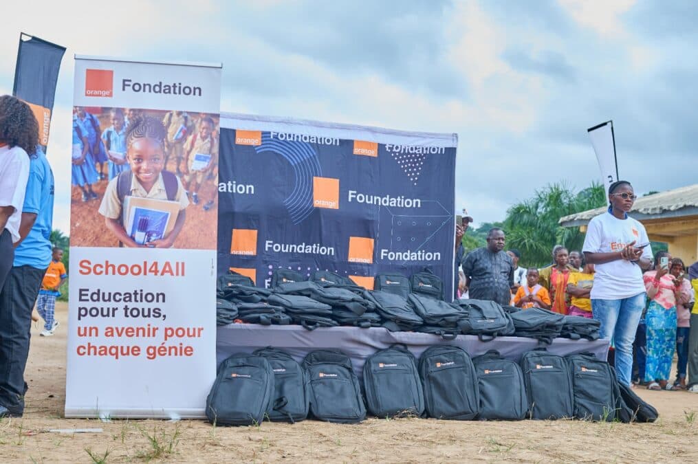 Une vue d'ensemble de la zone de distribution en extérieur. Au premier plan, des dizaines de cartables noirs sont alignés sur le sol et sur des tables couvertes d'un drap noir orné du logo de la fondation. À gauche, un grand kakemono affiche le message : « School4All. Éducation pour tous, un avenir pour chaque génie. » Quelques personnes, adultes et enfants, observent la scène en arrière-plan sous un ciel nuageux.