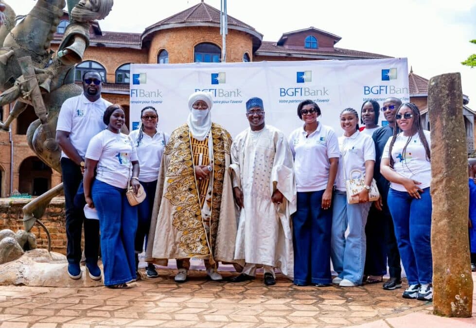Photo de groupe prise en extérieur devant la grande bannière BGFIBank et le Palais Royal de Foumban. Au centre figurent le Sultan des Bamoun et Abakal Mahamat, entourés des collaborateurs de la banque vêtus de t-shirts blancs aux couleurs de l'institution.