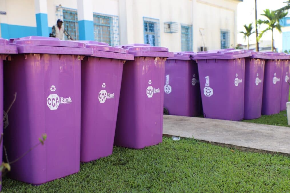 Alignement au premier plan de grands bacs à ordures violets de marque CCA-BANK, installés sur la pelouse extérieure de l'enceinte hospitalière de Laquintinie.