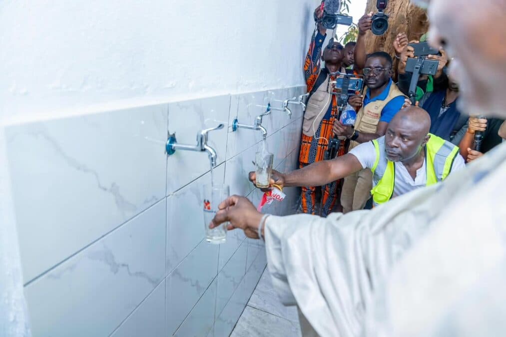 Vue rapprochée sur les robinets chromés fixés sur un mur carrelé de blanc. Deux verres sont remplis d'eau claire sous l'œil attentif des techniciens et sous les objectifs des caméras et photographes de presse.