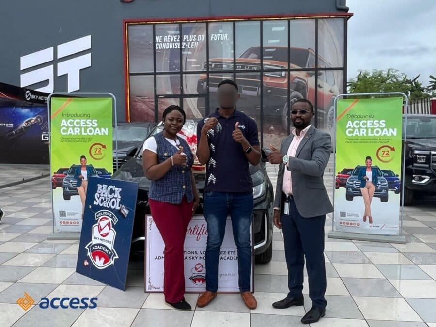 Une photographie en extérieur, en journée, sur le parvis carrelé du showroom Sky Motors à Douala, au Cameroun. Trois personnes posent pour la caméra, tout sourire, avec des pouces levés en signe de satisfaction. Au centre, l'homme (dont le visage est flouté pour la confidentialité) porte un polo bleu et tient fièrement une clé de voiture dans sa main droite et le certificat encadré de la Sky Motors Academy dans sa main gauche. À sa gauche, une dame représentant Sky Motors est vêtue d'un gilet en jean et d'un pantalon rouge. À sa droite, un homme en costume gris clair représente Access Bank Cameroon. Ils sont positionnés devant le capot d'un SUV Jetour X50 noir neuf, sur lequel repose un panier-cadeau emballé. De part et d'autre, des kakémonos publicitaires de couleur verte pour le produit « Access Car Loan » sont visibles, ainsi qu'un panneau de sol bleu portant l'inscription « BACK TO SCHOOL / WELCOME TO SKY MOTORS ACADEMY ». Le logo Access Bank est présent dans l'angle inférieur gauche de l'image.