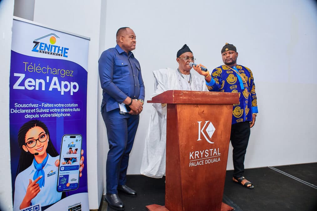 La scène se déroule sur une estrade devant un pupitre en bois portant le logo "K KRYSTAL PALACE DOUALA". Au centre, un homme d'un certain âge, vêtu d'une magnifique tenue traditionnelle camerounaise (blanche avec des broderies noires) et d'une coiffe assortie, parle dans un micro. À sa gauche, un homme en tenue de ville bleu marine observe attentivement. À sa droite, un autre homme porte une chemise en tissu traditionnel d'Afrique de l'Ouest (jaune et bleu) et une coiffe traditionnelle, tenant le micro. Un grand kakemono "Téléchargez Zen'App" avec un visuel de l'application et de son assistante virtuelle est visible sur la gauche. L'éclairage est professionnel et la scène est nette.